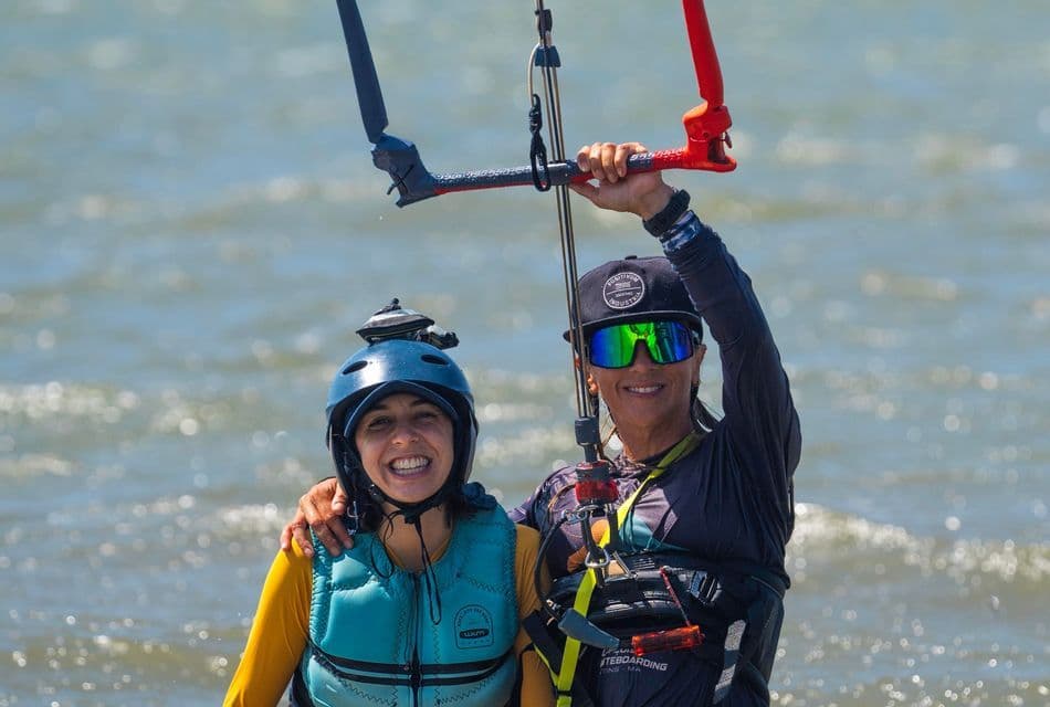 Due donne in attrezzatura per sport acquatici, inclusi caschi e giubbotti di salvataggio, sorridono nell'acqua mentre una tiene in mano una barra di controllo per kitesurf.