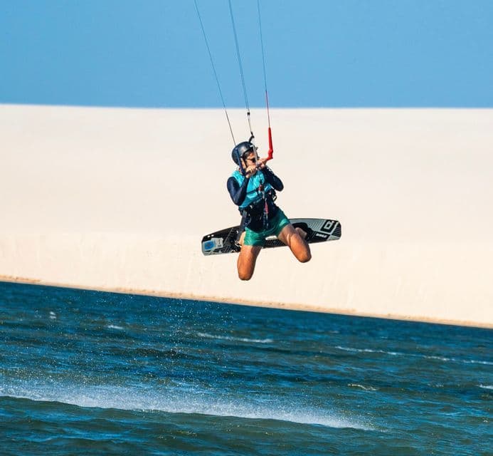 Una persona fa kitesurf librandosi sull'acqua blu, con una grande duna di sabbia bianca sullo sfondo.