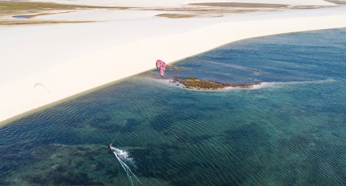 Vista aerea di una persona che fa kitesurf in acque turchesi vicino a una duna di sabbia bianca, con un aquilone rosa sulla riva.
