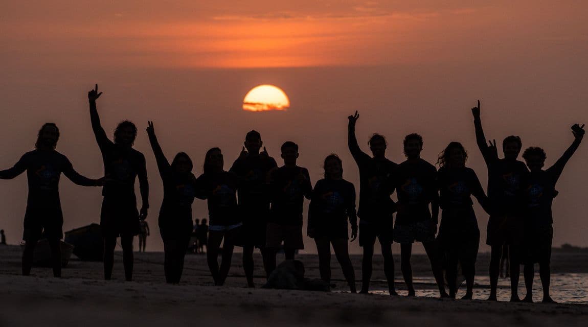 Un gruppo WeRoad in posa in silhouette su una spiaggia, con le braccia alzate contro il sole al tramonto e un cielo arancione.