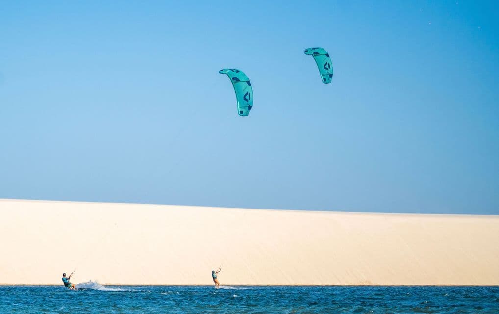 Due persone fanno kitesurf su acqua blu con grandi aquiloni verde acqua, con una duna di sabbia bianca sullo sfondo sotto un cielo sereno.