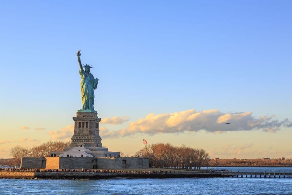 Die Freiheitsstatue thront auf ihrem Inselpodest und wird vom Wasser aus unter blauem Himmel mit Wolken bei Sonnenuntergang betrachtet.