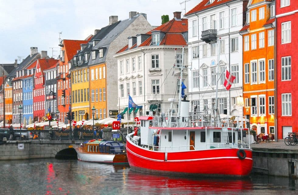 A red boat and other vessels are moored in a canal, with a row of colorful buildings lining the waterfront.