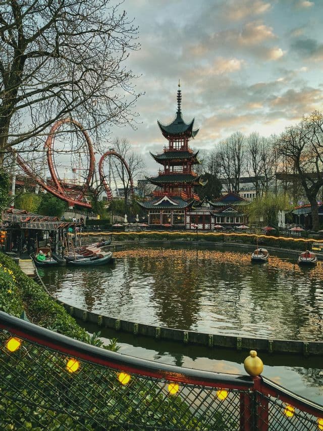 A tall pagoda stands beside a lake with boats in an amusement park, with a red rollercoaster in the background under a cloudy sky at dusk.