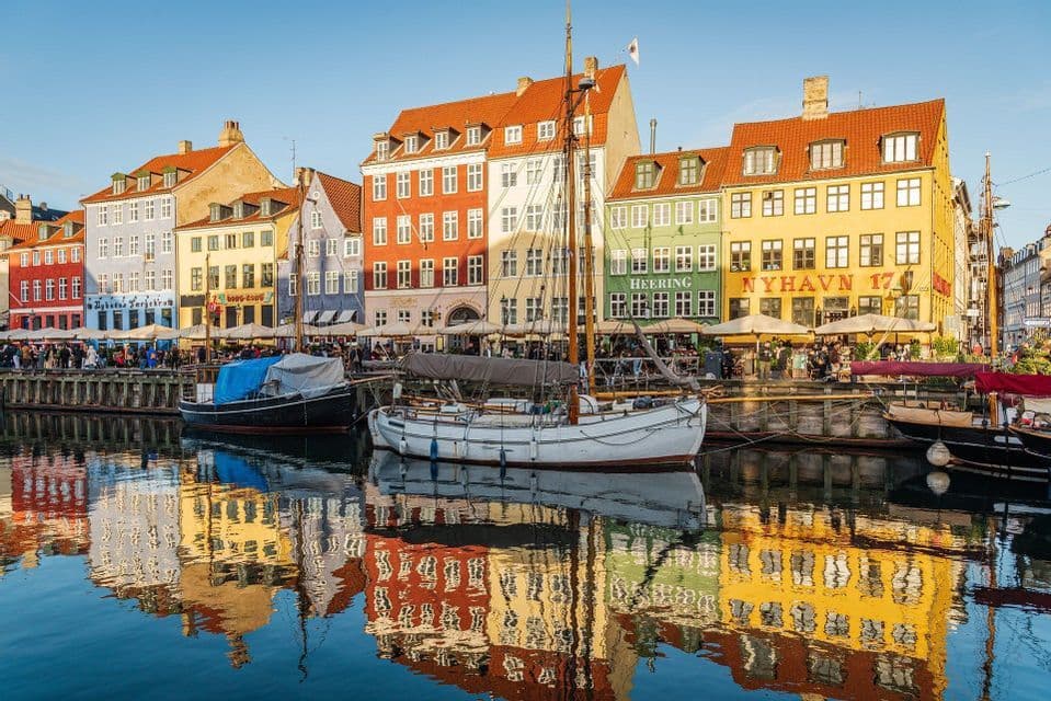 Colorful buildings and moored sailboats are reflected in the calm water of a canal under a clear blue sky.