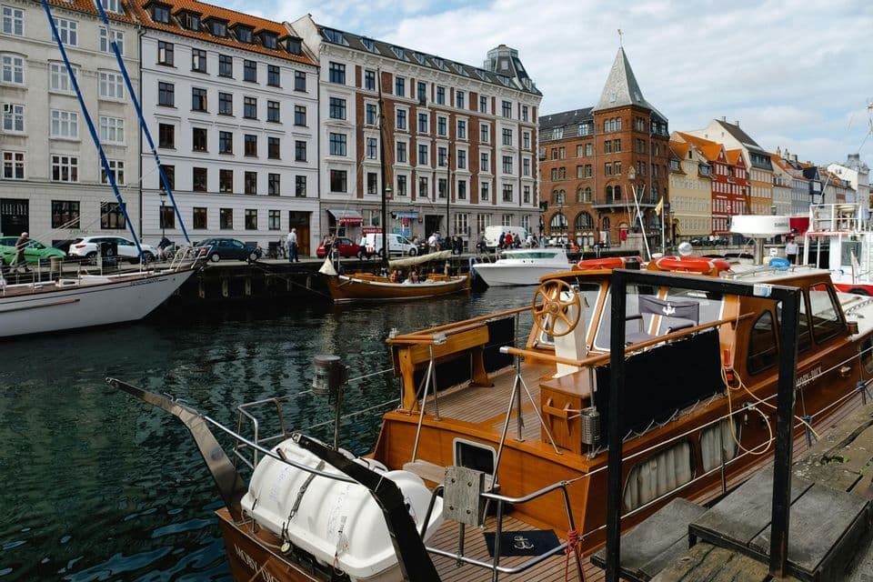 A wooden motorboat is docked in a city canal, with other boats and historic buildings lining the waterfront.