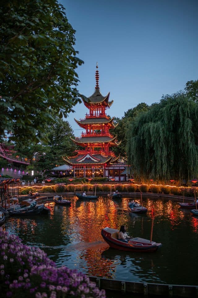 A large, red pagoda is illuminated at twilight, its lights reflecting on a pond where people ride in small boats.