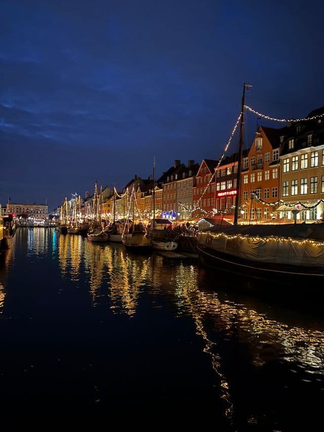 Boats and colorful buildings decorated with festive lights line a canal at night, their reflections shimmering on the water.