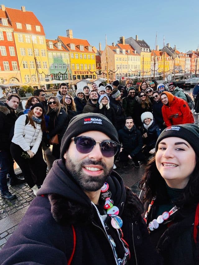 A WeRoad group trip poses for a selfie on a cobblestone street in front of colorful buildings along a harbor.