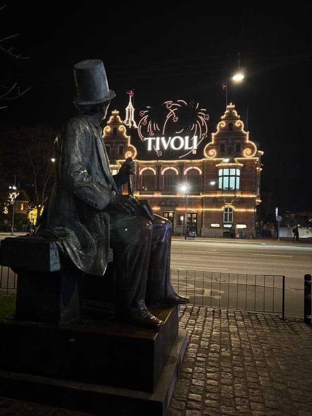 A bronze statue of a man wearing a top hat sits on a bench overlooking the illuminated Tivoli building at night.