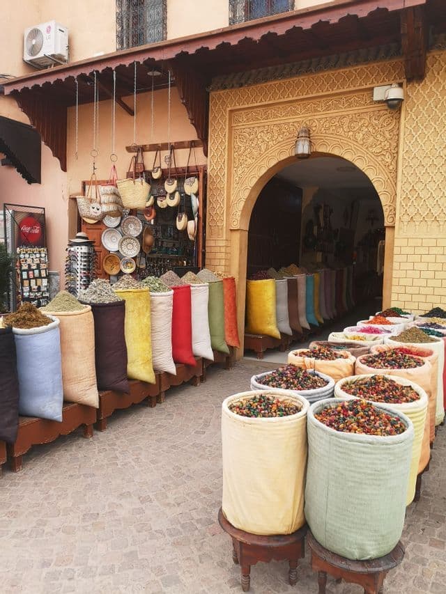 Bunte Säcke voller Gewürze und Potpourri, ausgestellt vor einem Marktladen mit einem reich verzierten Torbogen an einer Kopfsteinpflasterstraße.