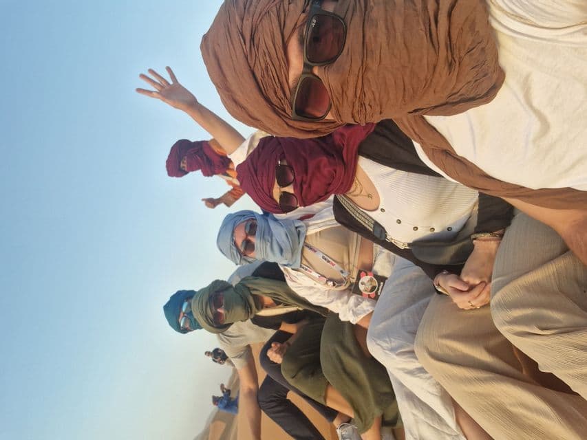 Eine WeRoad-Gruppe, die Sonnenbrillen und bunte Kopftücher trägt, posiert für ein Selfie aus der Froschperspektive vor klarem Himmel.