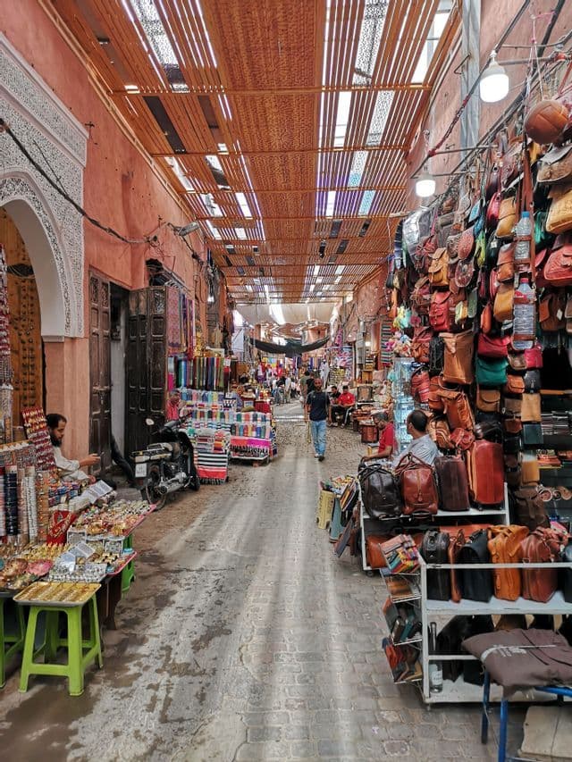 Eine belebte, enge Marktstraße mit Geschäften, die Ledertaschen und bunte Textilien unter einem Lamellendach anbieten.