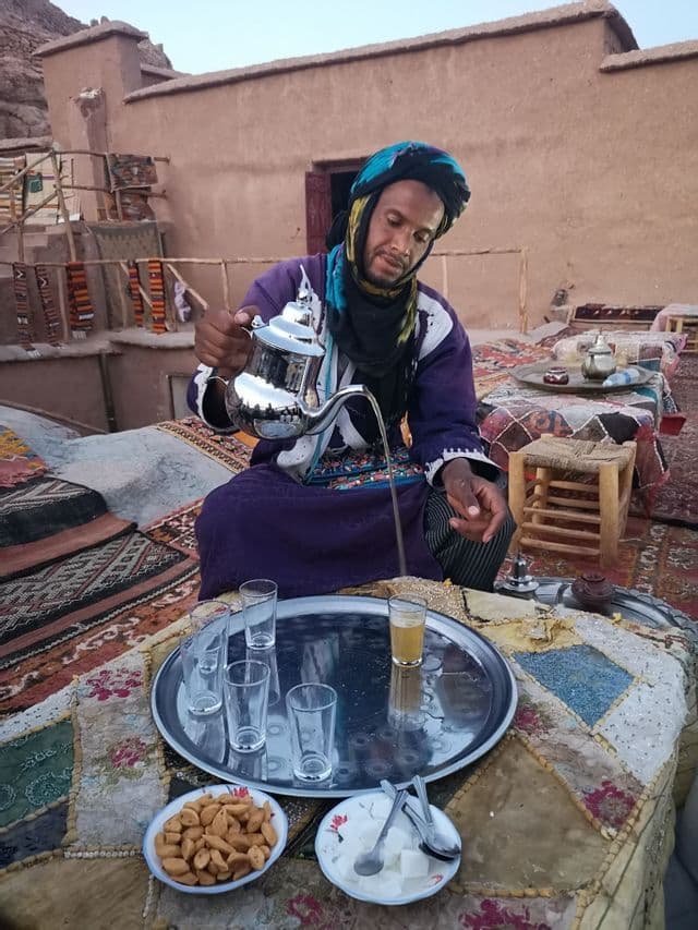 Ein Mann in einem violetten Gewand und blauem Turban gießt Tee aus einer silbernen Teekanne in ein Glas auf einem Tablett, sitzend auf farbenfrohen Teppichen.