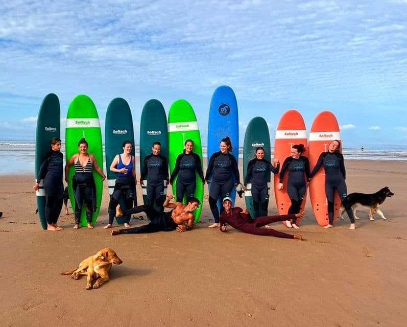 Eine WeRoad Gruppenreise in Neoprenanzügen posiert mit Surfbrettern und zwei Hunden an einem Sandstrand.