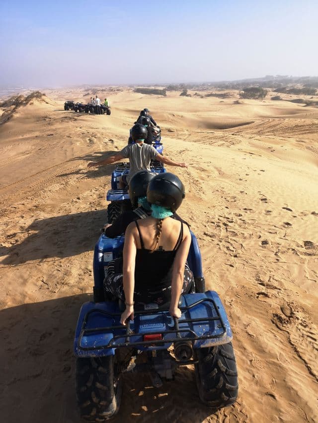 Eine WeRoad-Gruppe fährt mit blauen Quads in einer Reihe über Wüstensanddünen unter strahlendem Himmel.
