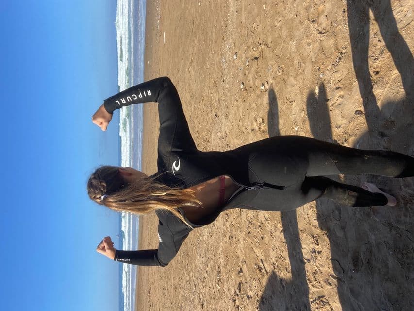 Eine Frau in einem schwarzen Neoprenanzug kniet an einem Sandstrand und spannt ihre Arme an, mit dem Ozean und blauem Himmel im Hintergrund.