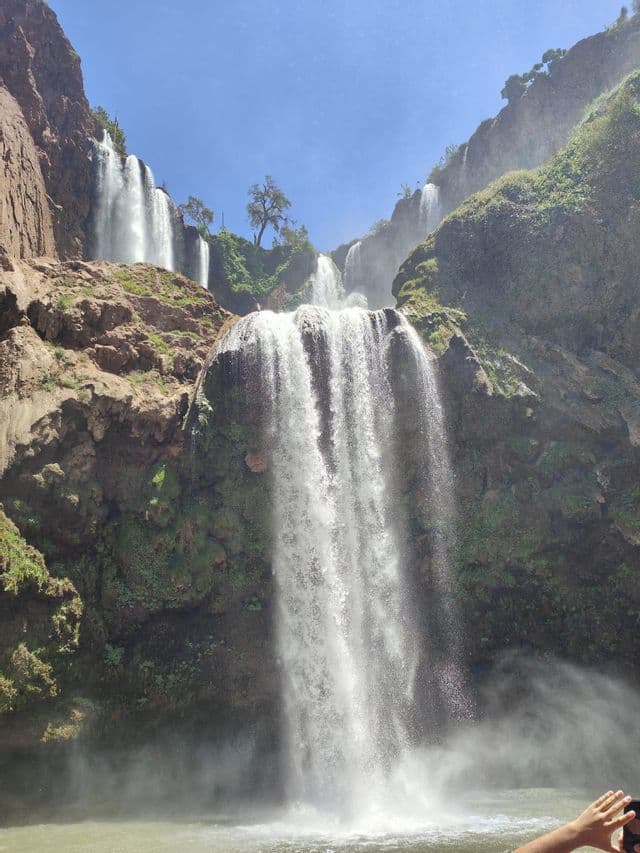 Mehrere Wasserfälle stürzen felsige, moosbewachsene Klippen hinunter in ein Wasserbecken unter strahlend blauem Himmel.
