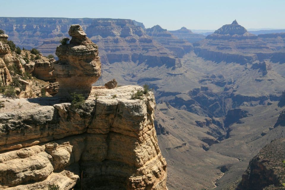 Una formación rocosa erosionada se asienta al borde de un acantilado, con vistas a la vasta extensión de un cañón escalonado bajo un cielo despejado.