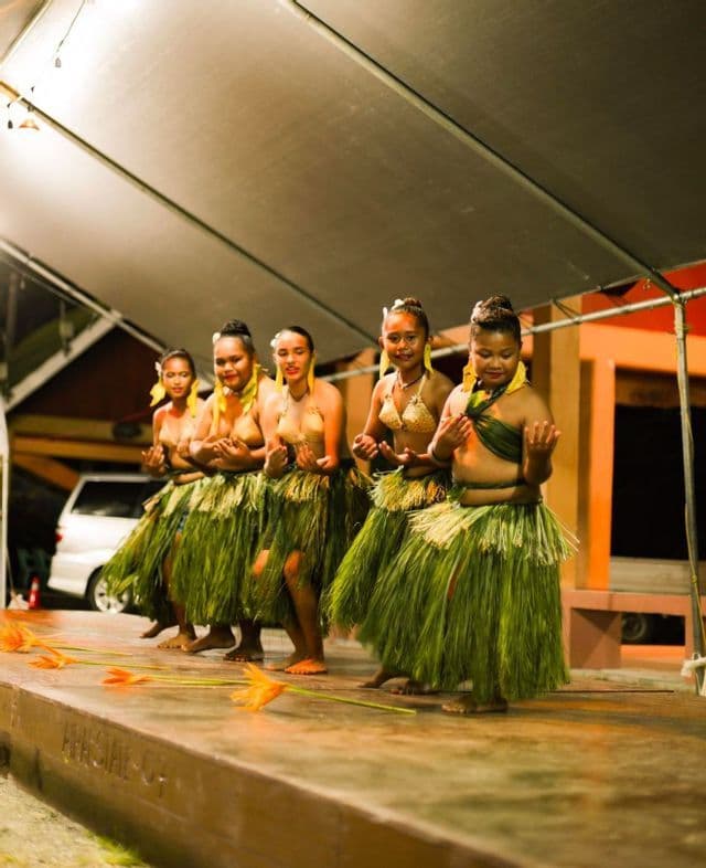 Un gruppo di ballerine con gonne tradizionali d'erba verde che si esibiscono su un palco di legno di notte.