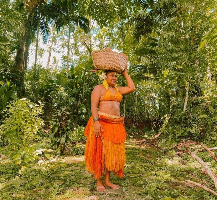 Una donna con una tradizionale gonna e top di erba arancione porta un cesto intrecciato sulla testa mentre si trova su un sentiero nella giungla.