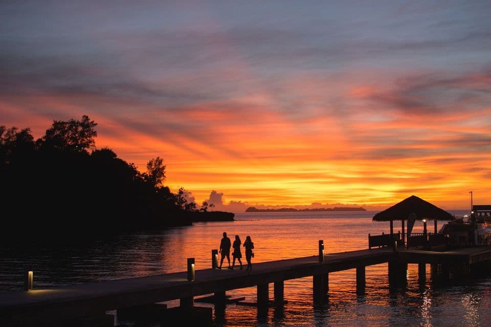 Un viaggio di gruppo WeRoad con tre persone in silhouette su un molo al tramonto, con un cielo arancione che si riflette sull'oceano.