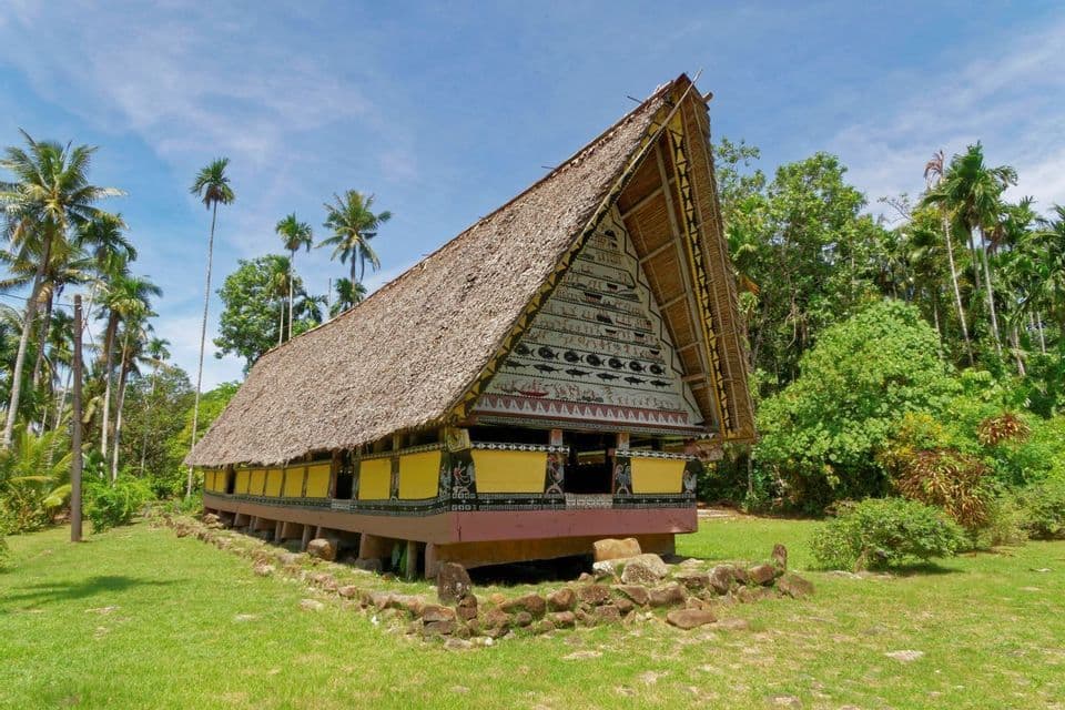 Un edificio tradizionale con un ripido tetto di paglia e una facciata dipinta sorge in una radura erbosa circondata da palme.