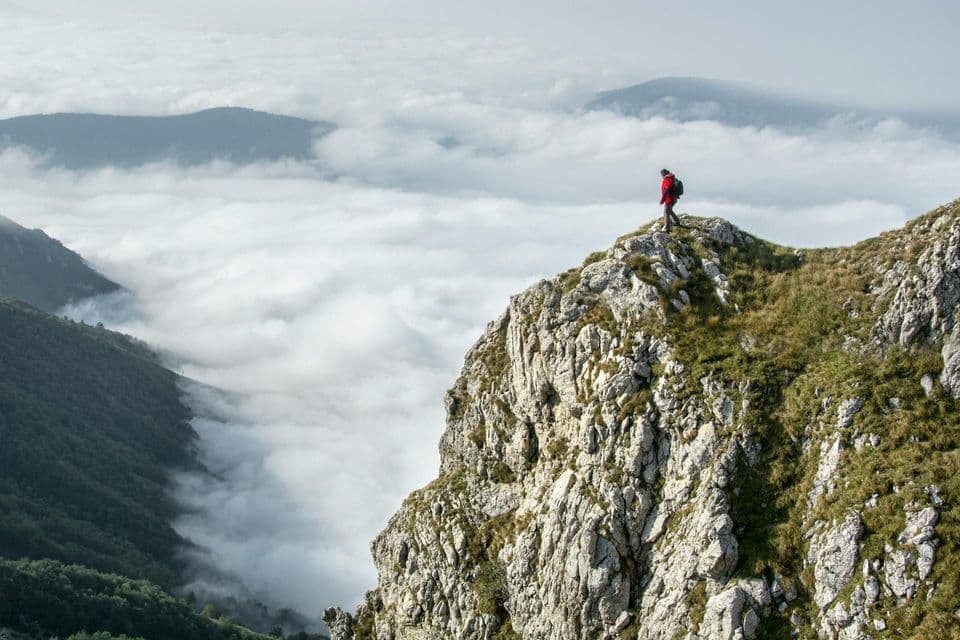 Ein Wanderer mit roter Jacke und Rucksack steht auf einem felsigen Berggipfel und blickt über ein Wolkenmeer.
