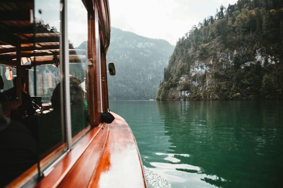 Ein Holzboot segelt auf einem grünen See neben steilen, bewaldeten Bergen.