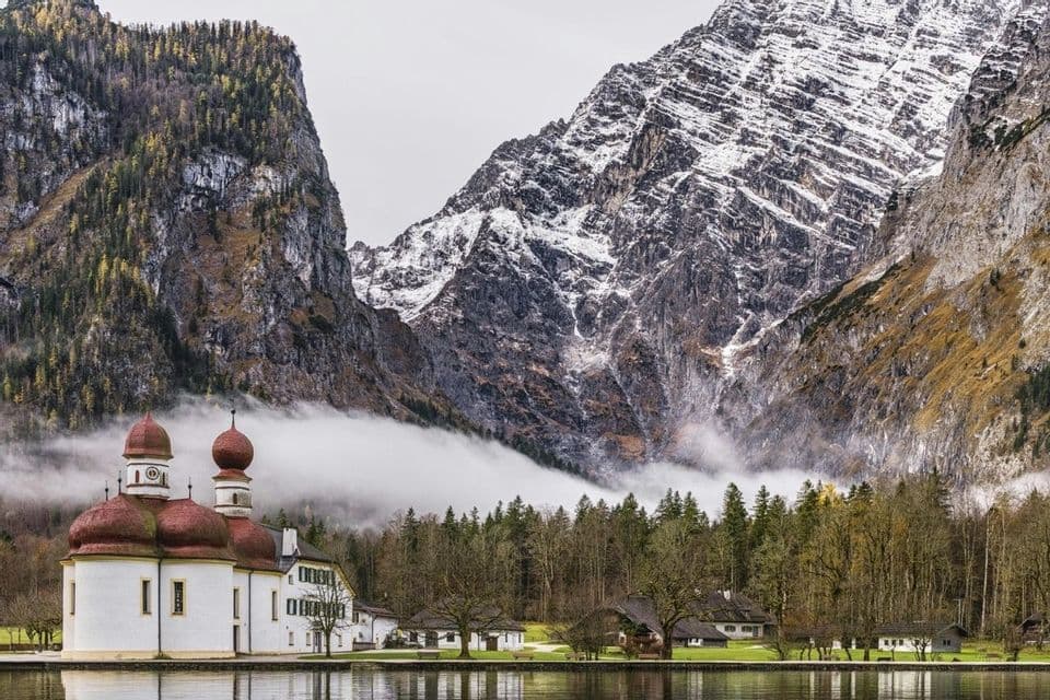 Eine weiße Kirche mit roten Zwiebelkuppeln steht am Seeufer, dahinter erheben sich hohe, schneebedeckte Berge durch eine Nebelschicht.