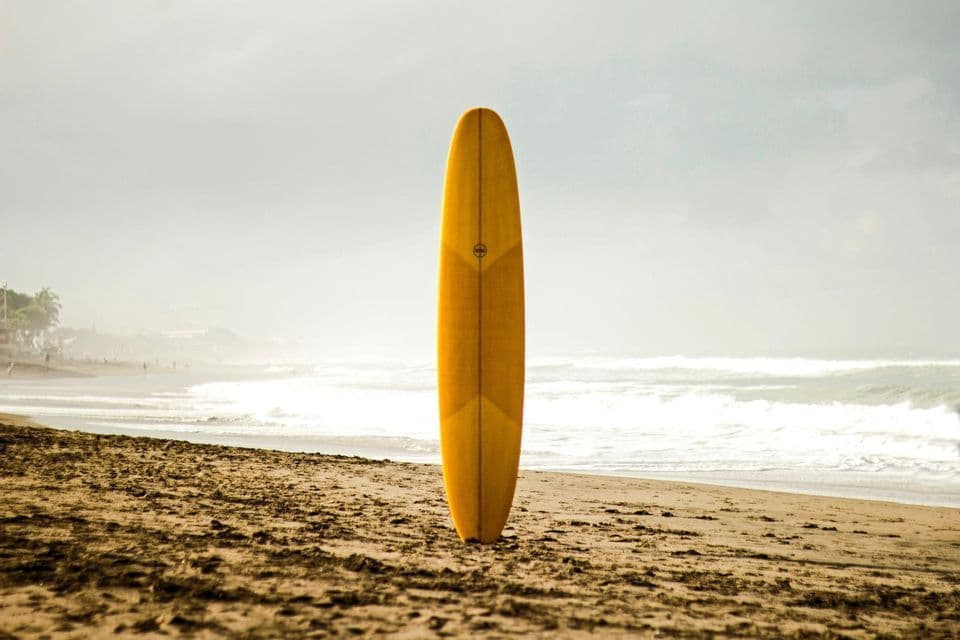 Una tabla de surf amarilla se mantiene erguida en la arena de una playa, con olas del océano y un cielo nublado de fondo.