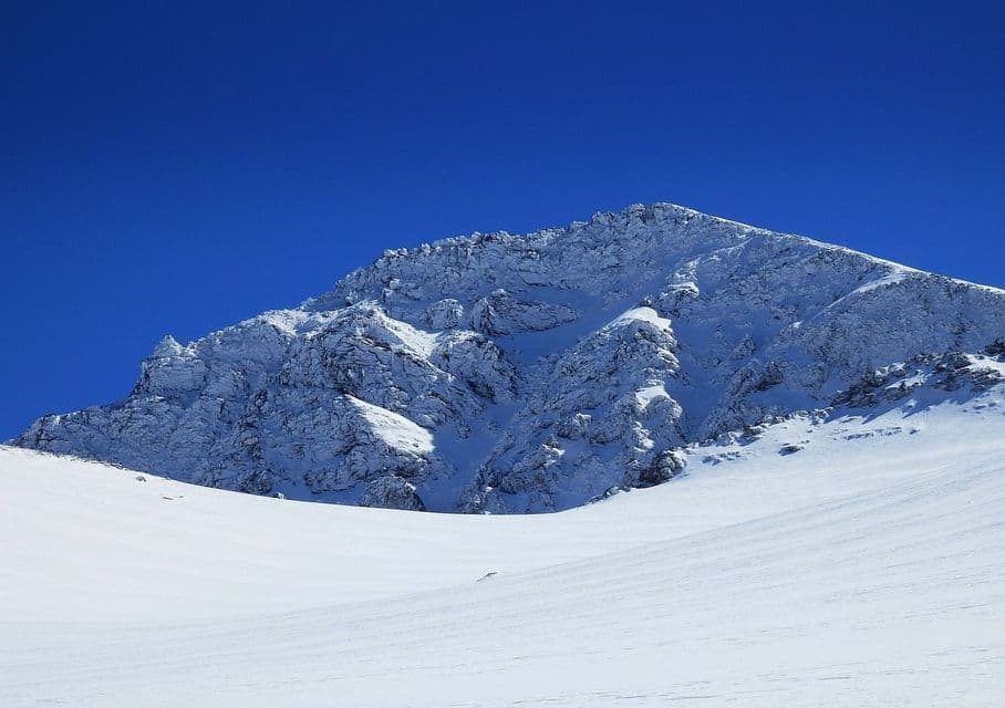 Un pico rocoso cubierto de nieve contrasta con un cielo azul profundo y sin nubes, sobre una vasta ladera nevada.