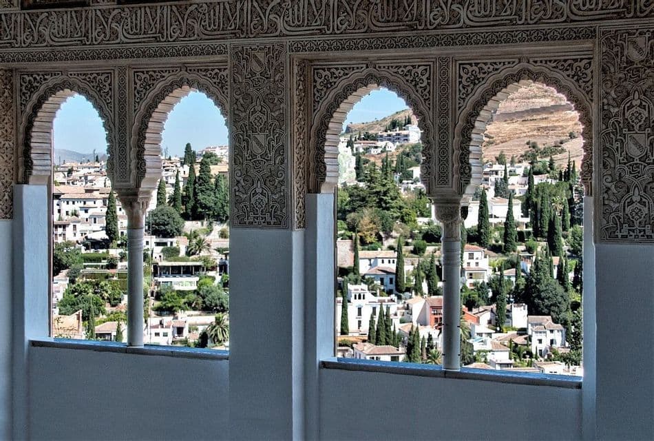 Un pueblo soleado en la ladera con edificios blancos y árboles verdes, visto a través de una serie de ventanas arqueadas moriscas, ornamentadas y festoneadas.