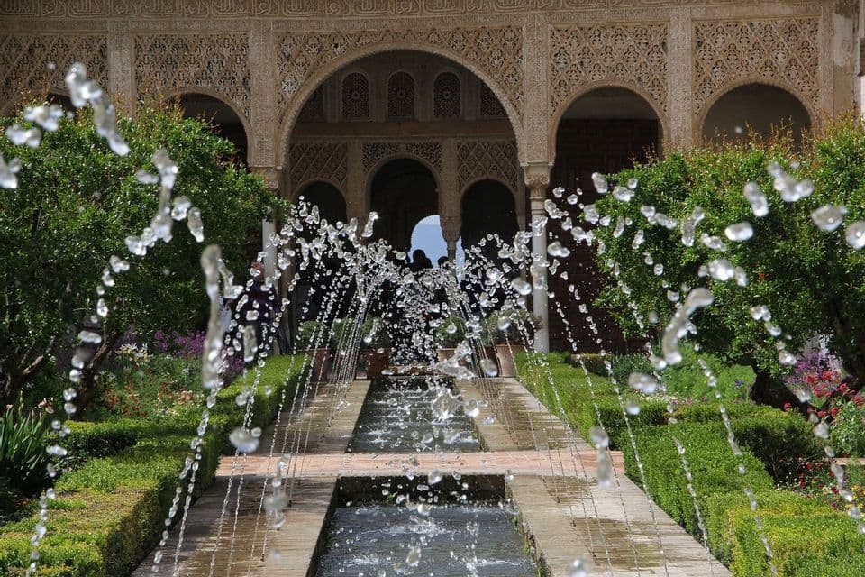 El agua brota de una larga fuente en un patio ajardinado, con arquitectura ornamentada y arqueada y arbustos verdes al fondo.
