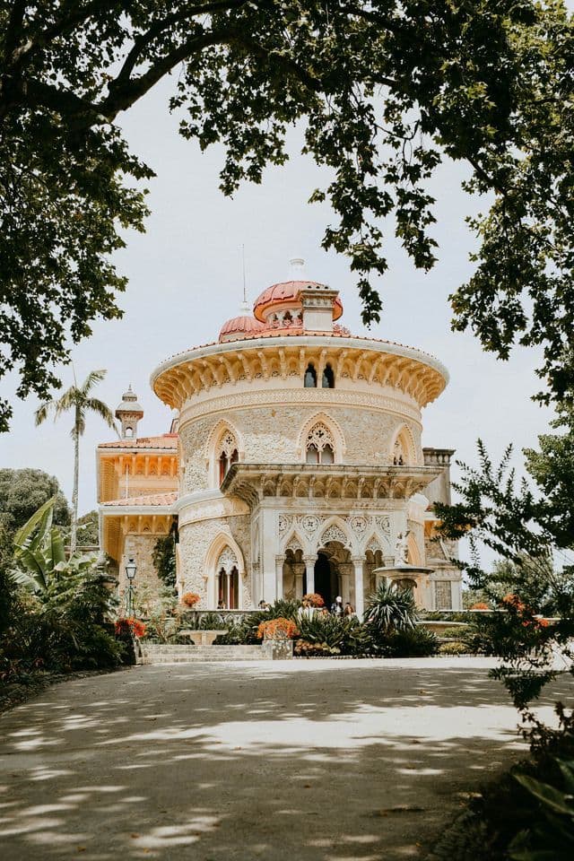 Un sontuoso palazzo circolare in pietra con cupola rossa, visto da un sentiero ombreggiato e incorniciato da foglie verdi.