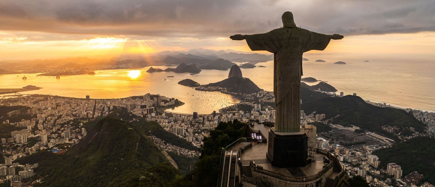 Eine große Statue mit weit ausgebreiteten Armen überragt bei Sonnenuntergang eine ausgedehnte Küstenstadt, eine Bucht und Berge.