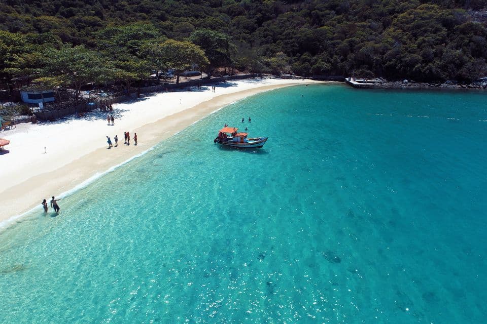 Vista aerea di una barca in acqua turchese vicino a una spiaggia di sabbia bianca dove le persone nuotano, con alle spalle una rigogliosa foresta verde.