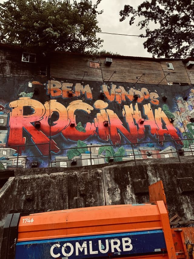 Un murale colorato di graffiti su un muro di cemento con la scritta 'Bem vindos Rocinha', con un camion arancione in primo piano.