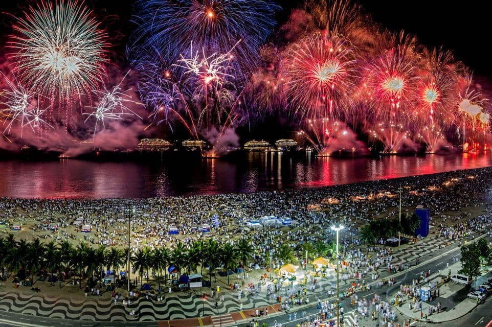 Farbenfrohes Feuerwerk explodiert am Nachthimmel über einer Bucht mit Schiffen, beobachtet von einer riesigen Menschenmenge an einem Sandstrand.