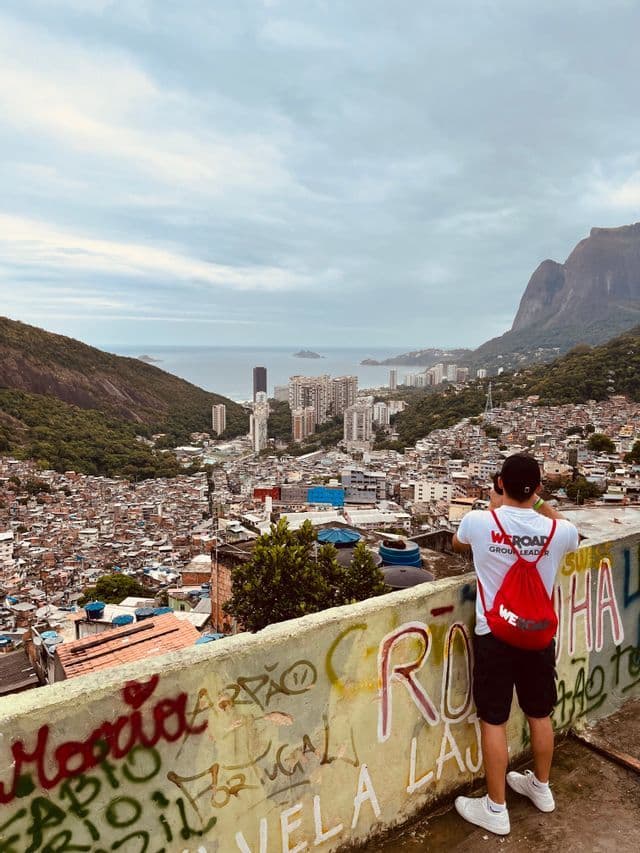 Ein WeRoad-Gruppenleiter mit rotem Rucksack fotografiert ein Favela-Stadtbild von einem Aussichtspunkt mit Blick auf das Meer.