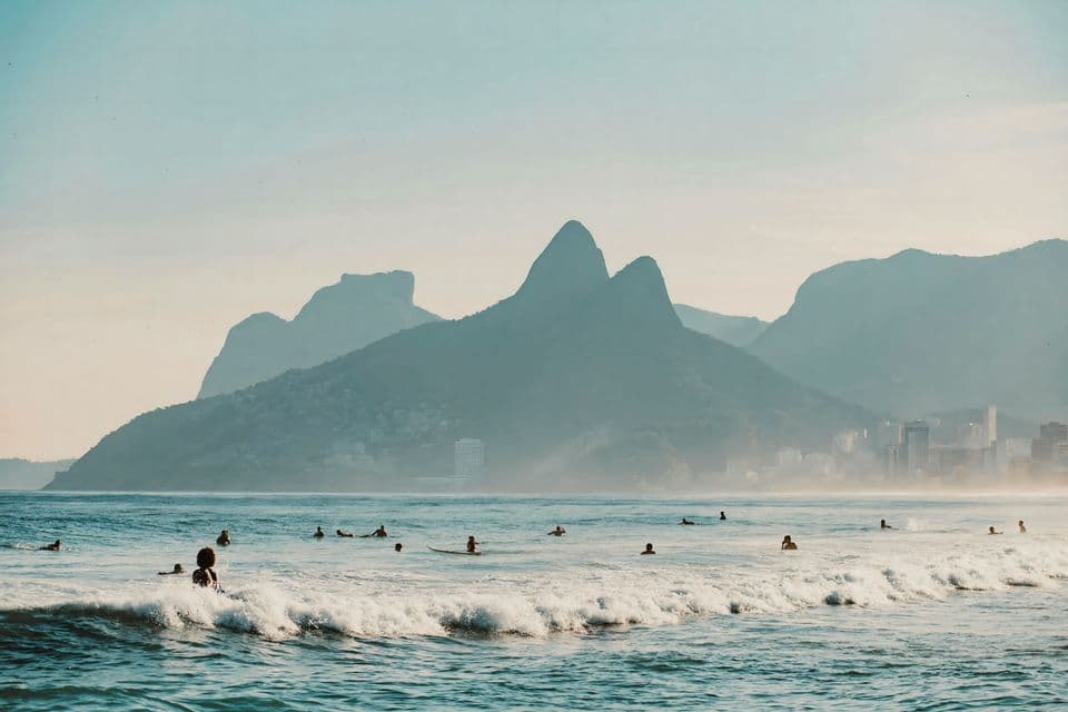 Un groupe de surfeurs attend les vagues dans l'océan, avec une chaîne de montagnes voilée et une ligne d'horizon urbaine en arrière-plan.