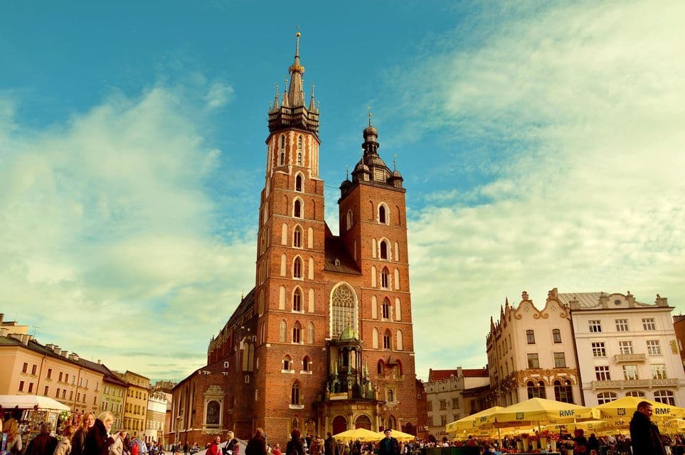 Eine Untersicht auf eine historische rote Backsteinkirche mit zwei Türmen auf einem belebten europäischen Platz unter blauem Himmel.