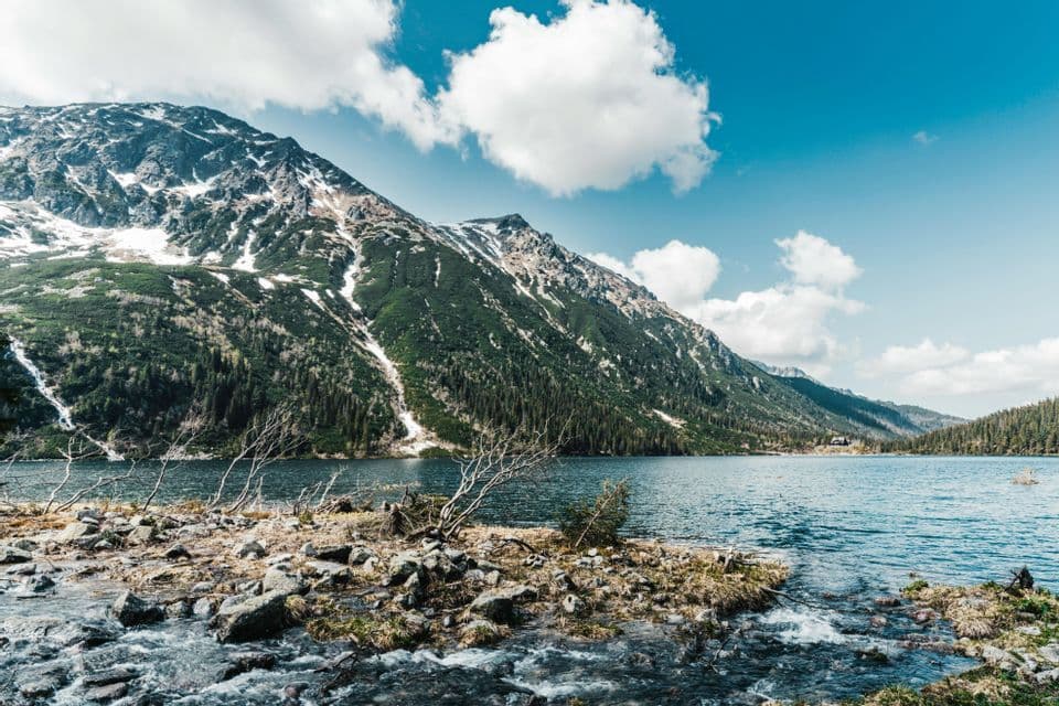 Ein breiter See liegt am Fuße eines großen, schneebedeckten und bewaldeten Berges, unter blauem Himmel mit Wolken.