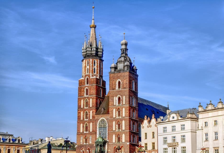 Zwei verzierte Backsteintürme einer historischen Kirche stehen vor einem klaren blauen Himmel neben anderen Gebäuden.