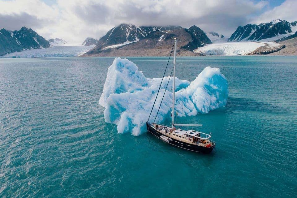 Un viaggio di gruppo WeRoad in barca a vela naviga accanto a un grande iceberg in acque artiche, con montagne e ghiacciai sullo sfondo.