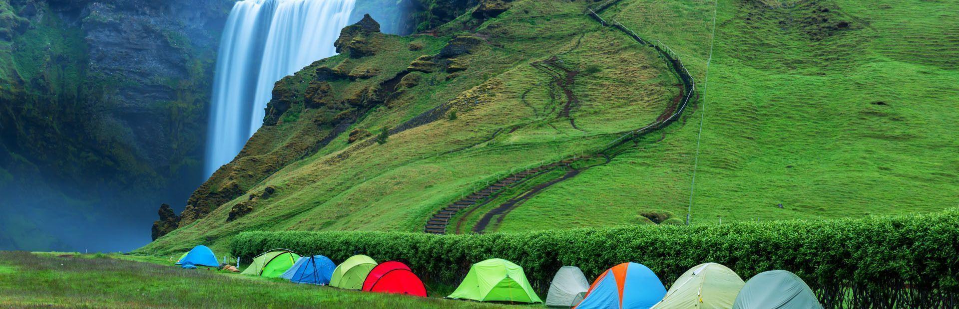 A row of colorful camping tents is lined up on a green field with a large waterfall and mossy cliff in the background.