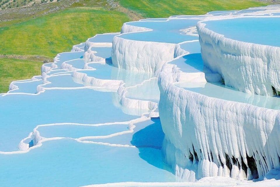 Una serie di terrazze bianche di travertino piene di acqua azzurra, con una collina verde visibile sullo sfondo.