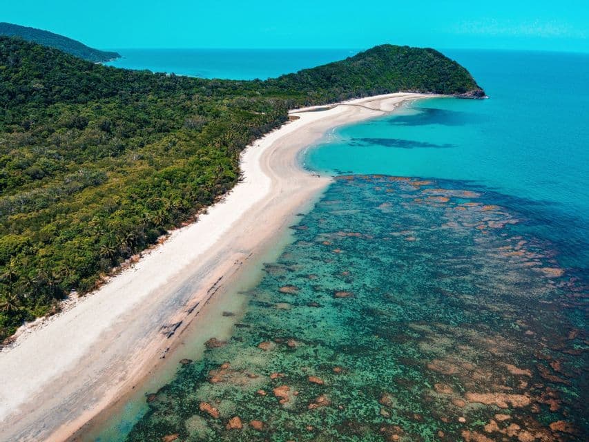 Vista aérea de una playa de arena blanca bordeando un exuberante bosque verde y un océano turquesa transparente que revela un arrecife de coral.