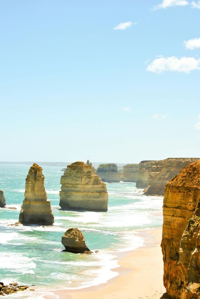 Grandes apilamientos de rocas se alzan en el océano turquesa junto a una playa de arena y acantilados bajo un cielo azul claro.