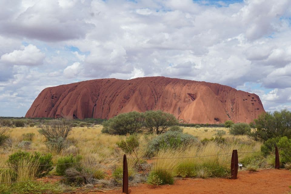 Un gran monolito de arenisca roja se alza desde una vasta llanura cubierta de hierba con arbustos dispersos bajo un cielo parcialmente nublado.
